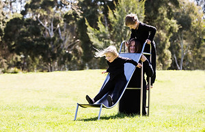 image: slope (2018), an artwork showing a series of images of a boy and a girl playing in the park on a three quarter sized wearable stainless steel slippery dip worn by their mother. It is a wearable neckpiece, with the mothers head poking through a hole in the top surface of the slide. The children are negotiating around the mothers head at the top of the ladder as they prepare to slide down © Tiffany Parbs, all rights reserved.