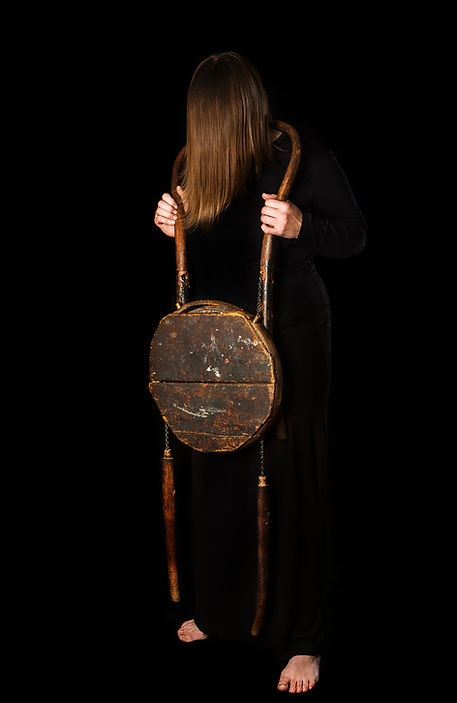 image: worn (2018), a jewellery artwork showing a woman whose face is obscured by her hair, dressed completely in black on a black background. She is wearing a necklace made from a deconstructed battered antique Brentwood chair. The deconstructed chair parts are held together with a linked chain, the curve of back of the chair rests on her neck and the seat of the chair is flat against her stomach with the front legs dangling down. The impression created is that she is feeling like part of the furniture © Tiffany Parbs, all rights reserved.
