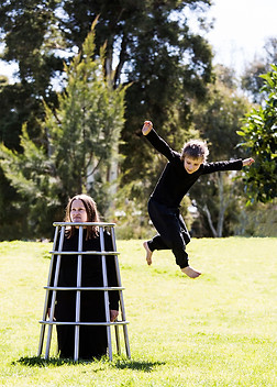 image: structure (2018), an artwork made of a series of images of two children playing in the park on a stainless steel climbing frame being worn by their kneeling mother. This image shows a boy jumping from the top stainless steel climbing frame being worn by his mother © Tiffany Parbs, all rights reserved.