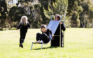 image: slope (2018), an artwork showing a series of images of a boy and a girl playing in the park on a three quarter sized wearable stainless steel slippery dip worn by their mother. It is a wearable neckpiece, with the mothers head poking through a hole in the top surface of the slide. The children are negotiating around the mothers head at the top of the ladder as they prepare to slide down © Tiffany Parbs, all rights reserved.