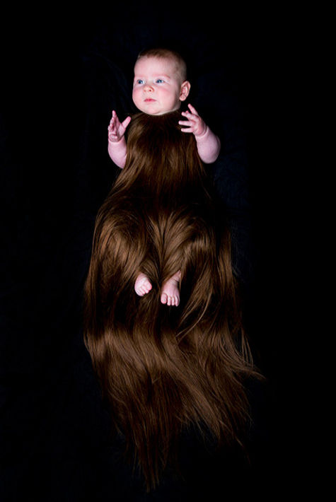 image: hirsute (2015), an artwork showing a red haired and fair skinned baby wearing a sleeveless onesie made from long auburn hair that cascades from the shoulders down past the feet. The baby appears swathed in the hair and suspended in front of a black background © Tiffany Parbs, all rights reserved.
