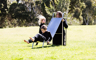 image: slope (2018), an artwork showing a series of images of a boy and a girl playing in the park on a three quarter sized wearable stainless steel slippery dip worn by their mother. It is a wearable neckpiece, with the mothers head poking through a hole in the top surface of the slide. The children are negotiating around the mothers head at the top of the ladder as they prepare to slide down © Tiffany Parbs, all rights reserved.