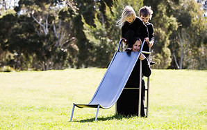 image: slope (2018), an artwork showing a series of images of a boy and a girl playing in the park on a three quarter sized wearable stainless steel slippery dip worn by their mother. It is a wearable neckpiece, with the mothers head poking through a hole in the top surface of the slide. The children are negotiating around the mothers head at the top of the ladder as they prepare to slide down © Tiffany Parbs, all rights reserved.