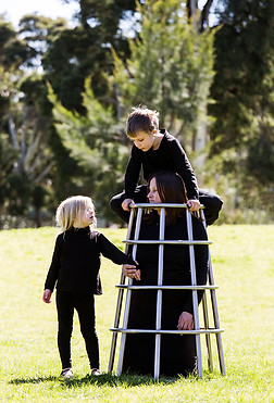 image: structure (2018), an artwork made of a series of images of two children playing in the park on a stainless steel climbing frame being worn by their kneeling mother. In this image the girl is looking into her mother face while the mother kneels inside the stainless steel cage and the boy is sitting on top looking down at his mothers head between his knees © Tiffany Parbs, all rights reserved.