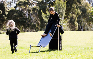 image: slope (2018), an artwork showing a series of images of a boy and a girl playing in the park on a three quarter sized wearable stainless steel slippery dip worn by their mother. It is a wearable neckpiece, with the mothers head poking through a hole in the top surface of the slide. The children are negotiating around the mothers head at the top of the ladder as they prepare to slide down © Tiffany Parbs, all rights reserved.