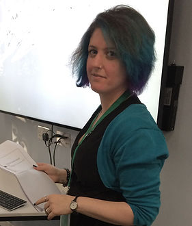 Louise, a woman in her 40s with a dark dod, looks at the camra. She is holding some notes, and behind her is a projector screen