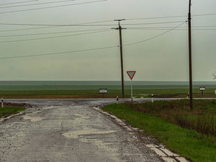 Camino rural mojado con baches, cercos y campos verdes al fondo.