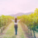 A photograph of a woman standing in a vineyard line, shown with her back to the camera and her arms outstretched as if to take in the world.