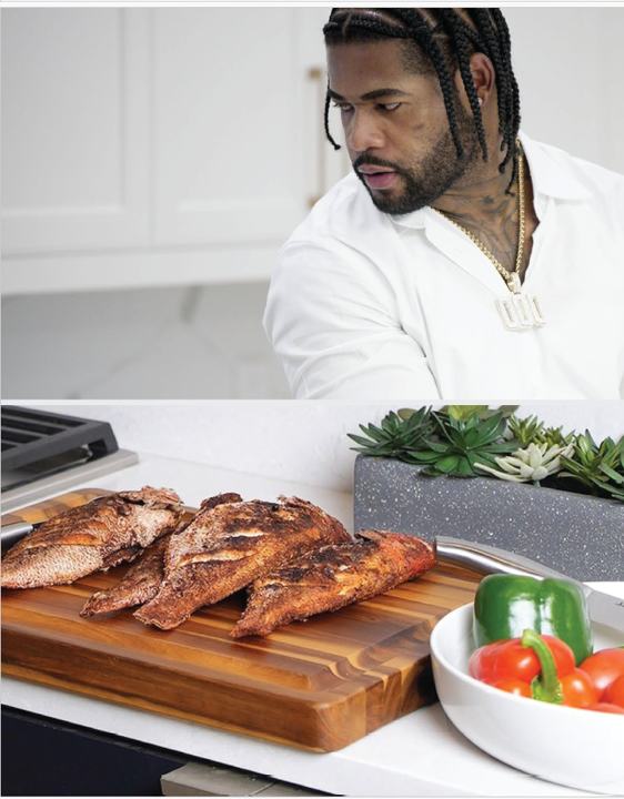 Chef O’Shane standing in the kitchen preparing Jamaican fried fish on a wooden cutting board, with fresh peppers and tomatoes beside the dish.