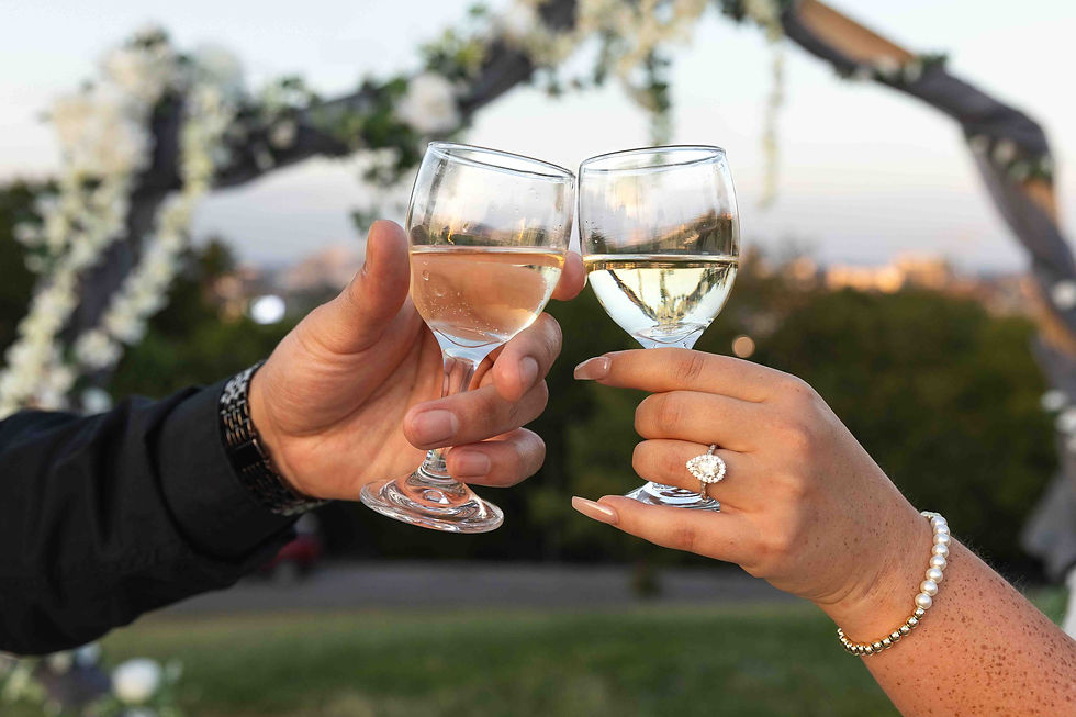 Two hands toast with wine glasses outdoors, one wearing a pearl bracelet and ring. A floral arch and greenery are blurred in the background.