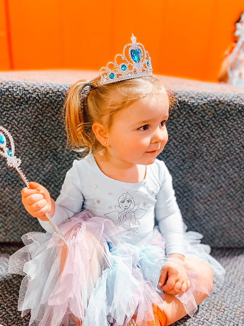 A young girl dressed as a princess, with a sparkling tiara and a beautiful gown, standing in a trampoline park specially decorated for a princess-themed event. She's eagerly awaiting the chance to meet a real princess, and her excitement is palpable in her radiant smile and shining eyes