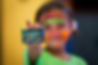 A photo of a young toddler girl at an indoor trampoline park with neon face paint, smiling at the camera while holding a membership card up.