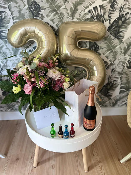 Feier zum 25-jährigen Jubiläum von der Pflegedienstleitung der Hauskrankenpflege Kossian: Goldene Ballons mit der Zahl 25, ein Blumenstrauß, eine Sektflasche und kleine Geschenke auf einem stilvollen runden Tisch vor einer tropischen Tapete.