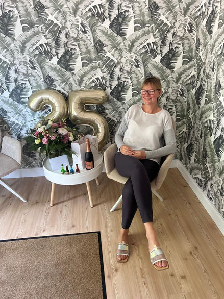 Jubiläumsfeier: Eine lächelnde Frau sitzt entspannt in einem modernen Raum mit tropischer Tapete. Neben ihr stehen goldene Luftballons mit der Zahl 25, ein Blumenstrauß und eine Flasche Sekt – ein festlicher Moment der Wertschätzung