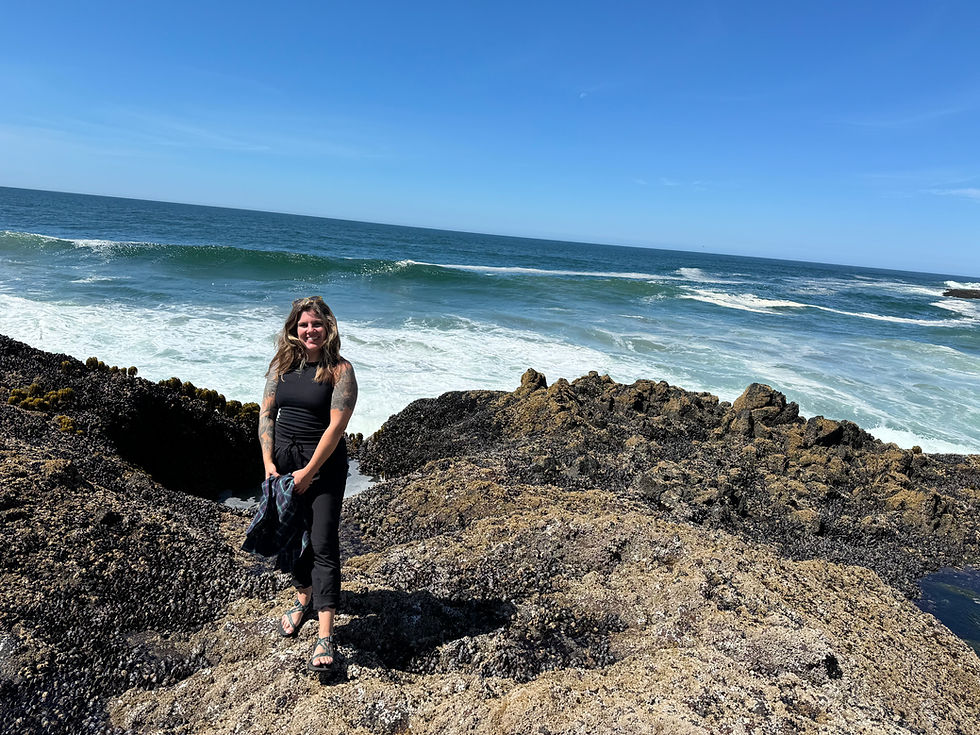 Pacific Ocean as seen from the Oregon coastline during my 2024 visit. (Image description: A woman with brown hair stands on large, dark rock formations at a beach, wearing a black tank top and dark pants while smiling at the camera. Behind her stretches a beautiful Pacific coastline with rolling waves and clear blue sky.)