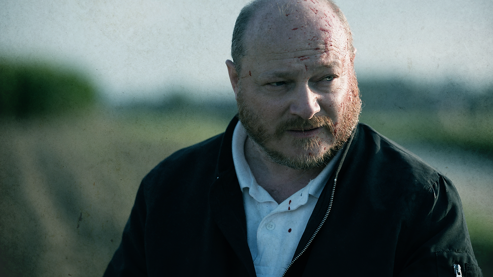 An image of actor Olivier Picard as the character Scola in the movie heat of Madness. In in the image, he's standing in a field, which is blurred in the background, with blood on his face and forehead.