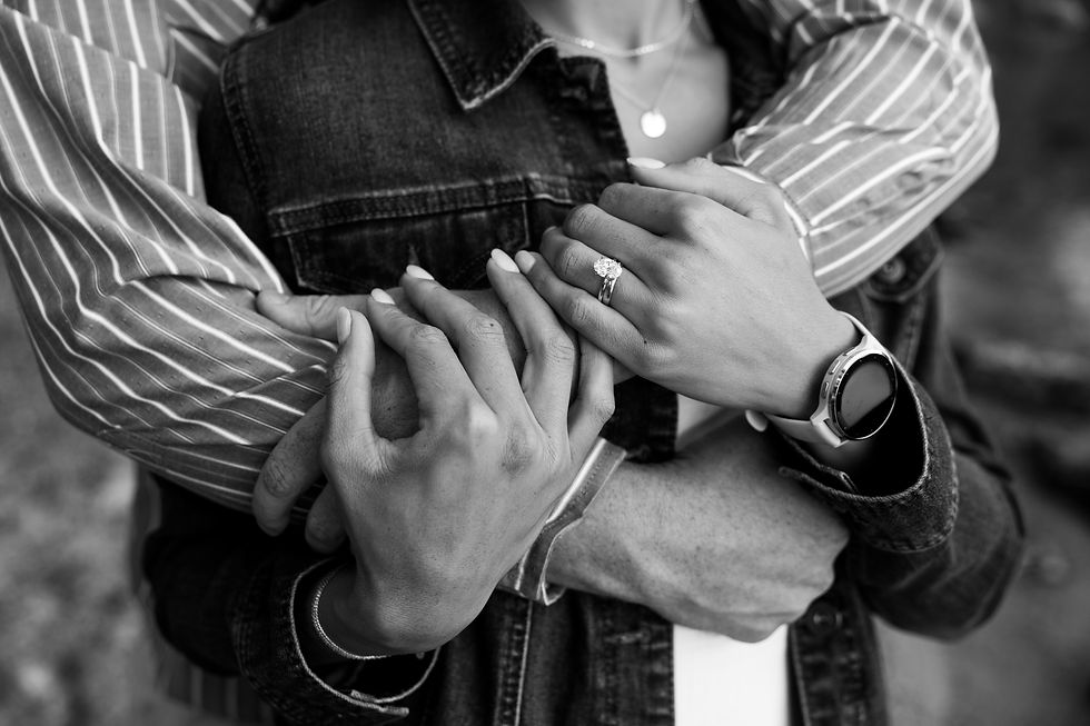 Close-up of a couple embracing, hands interlocked. Focus on a diamond ring and watch. Striped shirt and denim jacket visible. Black and white.