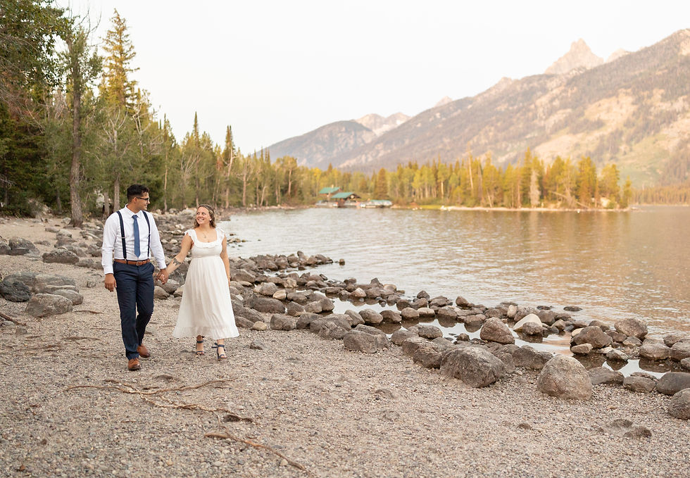 A couple walks hand in hand along a rocky lakeshore with mountains in the background. They appear happy, surrounded by serene nature.