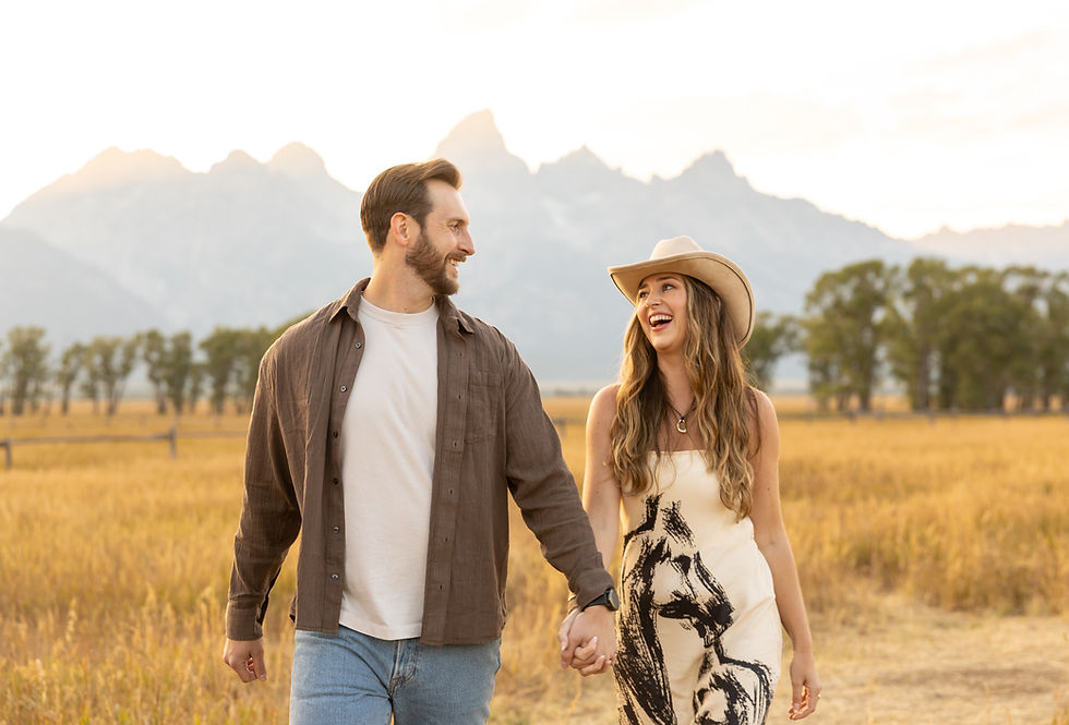 A couple holds hands and smiles at each other as they walk through a field with the grand teton mountains in the background.