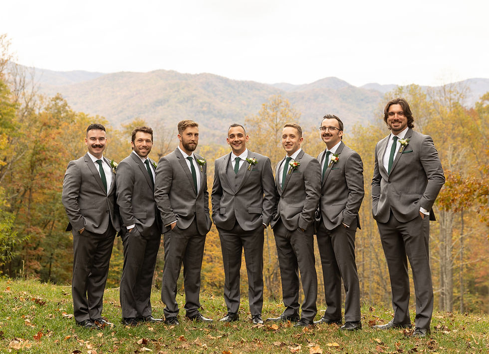 Groom and groomsmen in grey suits with green ties pose on grassy field with autumn foliage and mountains in background, looking cheerful.
