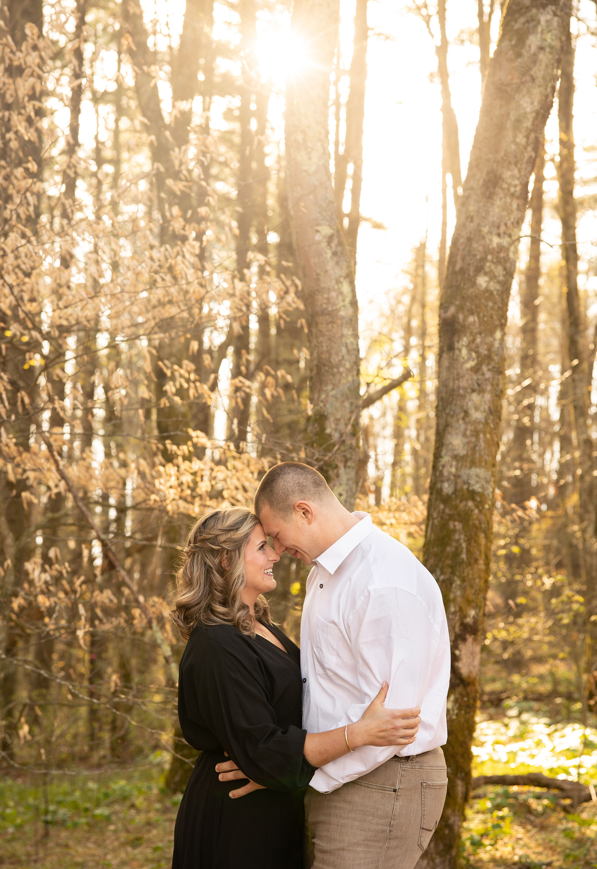 An engaged couples embraces each other close as the sun shines behind the trees.