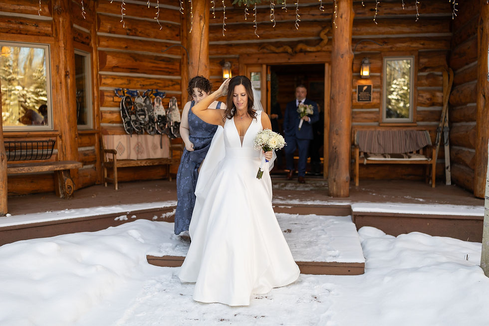 Bride in white gown holding bouquet, walking on snowy porch of a log cabin with bridesmaid adjusting dress. Groom in background with flowers.