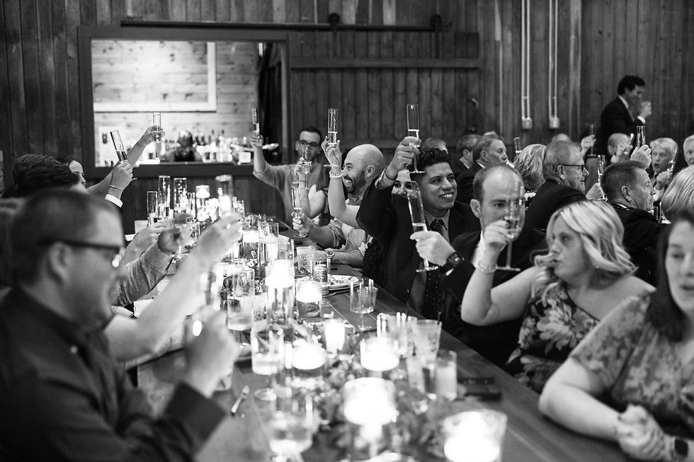 Wedding guests sitting at a long farm table and raising their glasses. It's in black and white and mood is cheerful.