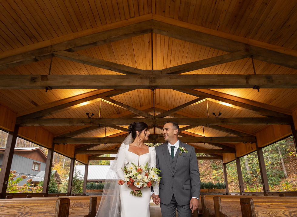 Bride and groom smiling under a wooden pavilion. The bride holds a colorful bouquet. Warm lighting and autumn foliage create a cozy atmosphere.
