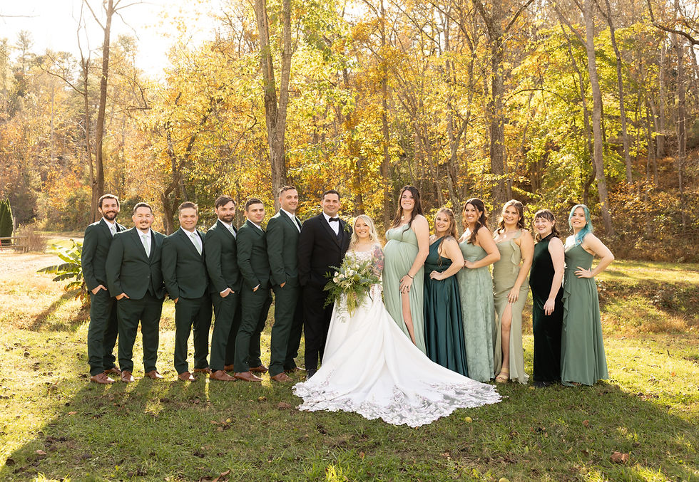 Wedding party poses outdoors in a sunlit forest. Bride in white dress holds bouquet, surrounded by groom in a tuxedo and attendants in green attire.