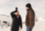 Two people in winter attire on a snowy mountain. One drinks from a large bottle, while the other looks on, both smiling under a clear sky.