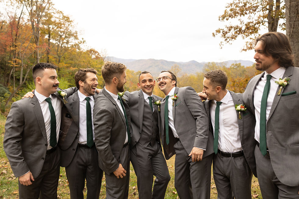 Seven men in matching gray suits and green ties laugh together outdoors, surrounded by fall trees and mountains, exuding joy and camaraderie.