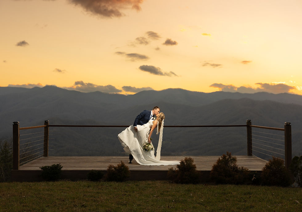 Bride and groom share a romantic kiss on a wooden deck at sunset, with mountains silhouetted in the background. Bride holds a bouquet.