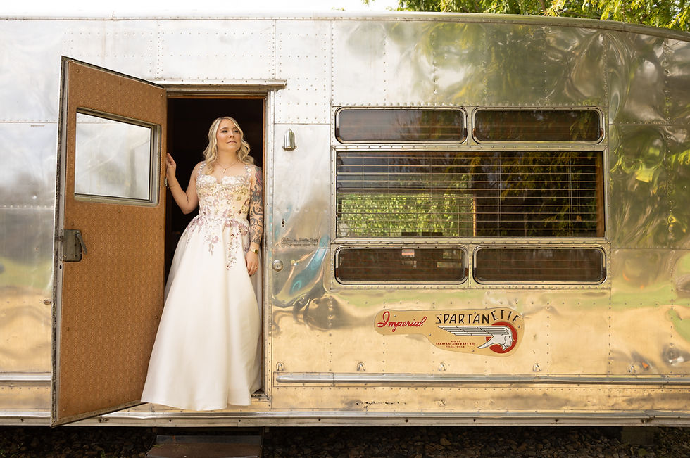 A woman in a white floral wedding dress stands at the open door of a shiny vintage trailer labeled "Imperial Spartanette," looking content.