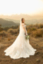 Bride in a lace gown smiles softly, holding flowers on a rocky hill. Sunlit mountains in the background create a serene, golden glow.