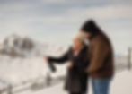 Woman joyfully sprays champagne, embraced by man in snowy mountain setting. Both in winter attire, with a clear sky and distant peaks.