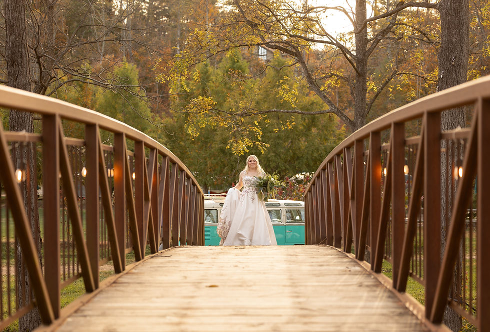 Bride in white dress with bouquet crosses wooden bridge in autumn setting. Vintage turquoise van in background; warm, serene mood.