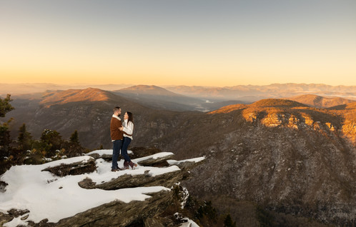 A couple stands together on a snow covered cliff in the mountains. 