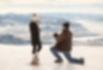Man proposes on snowy mountain, kneeling with ring box. Woman in winter attire looks surprised. Majestic snow-covered peaks in background.