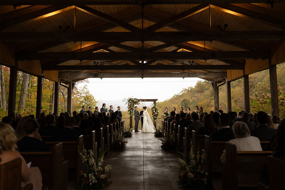 Bride and groom stand under a floral altar in a wooden pavilion, surrounded by guests and fall foliage, during an outdoor wedding ceremony.