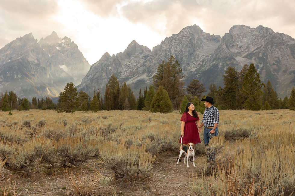 An engaged couple holds hands and walks their dog through an open field in front of the grand tetons. 