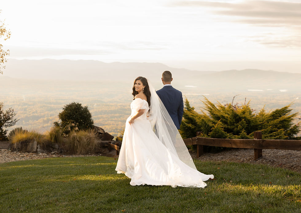 Bride in white gown and veil stands next to groom in blue suit on a grassy hilltop, with a scenic mountain view under a bright sky.