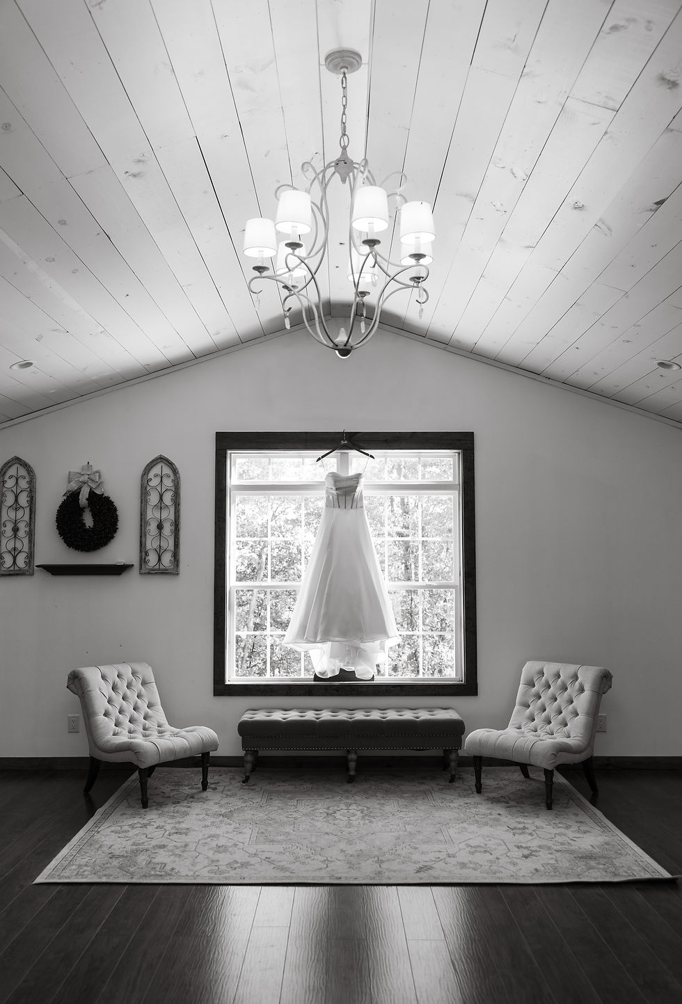 Wedding dress hangs near window in elegant room with white ceiling, chandelier, two tufted chairs, and a wreath on the wall.