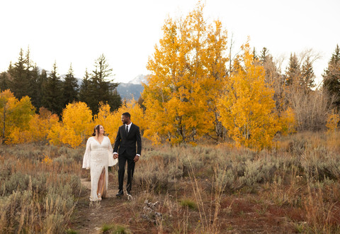 A bride and groom looking at each other intimately as they hold hands and walk across a field with yellow aspen trees in the background.