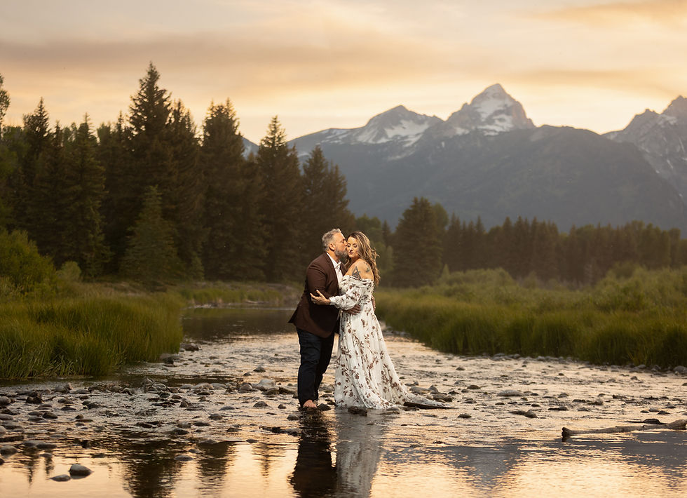 A couple embraces in a stream, surrounded by greenery and mountains. The sky is orange at sunset, creating a romantic mood. They are wearing non-traditional wedding attire.