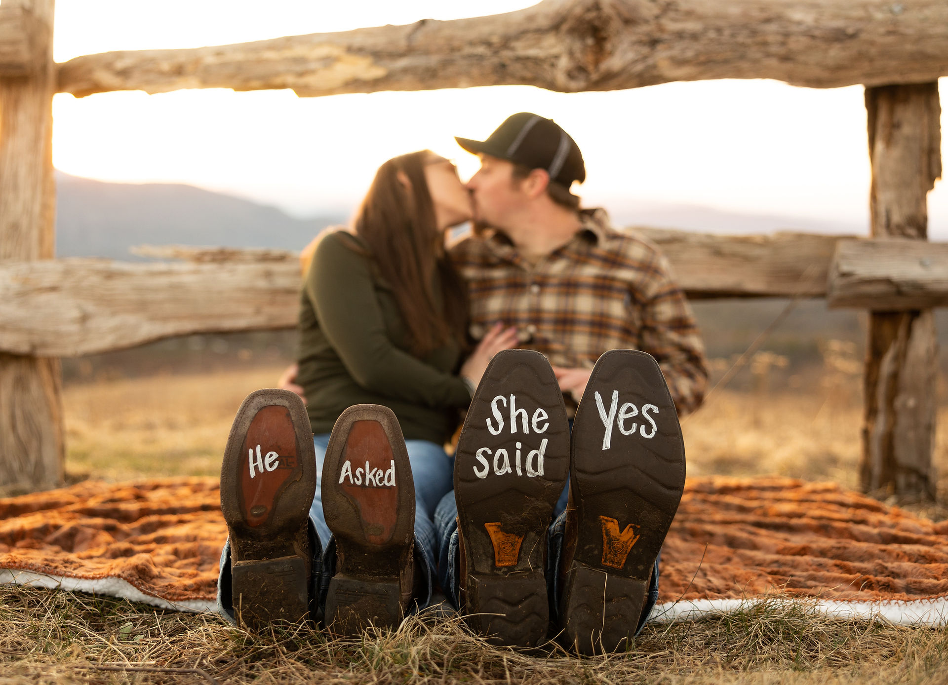 A young couple sits together leaning against a fence while the words he ask, she said yes is written on the bottom of their shoes.