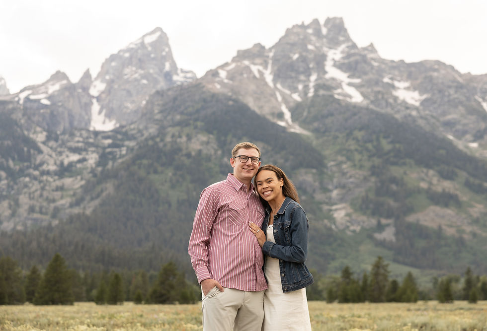 Couple smiling and embracing in a meadow with snow-capped mountains in the background. The man wears glasses and a striped shirt; the woman wears a denim jacket.