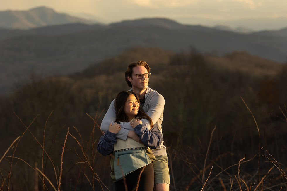 A couple embraces with smiles in a field at sunset, surrounded by mountains. The scene conveys warmth and tranquility.