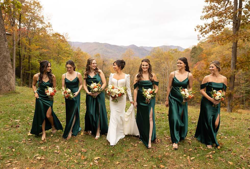Bride in white and bridesmaids in green dresses walk on grass, holding bouquets. Autumn trees and mountains in the background. Smiling.