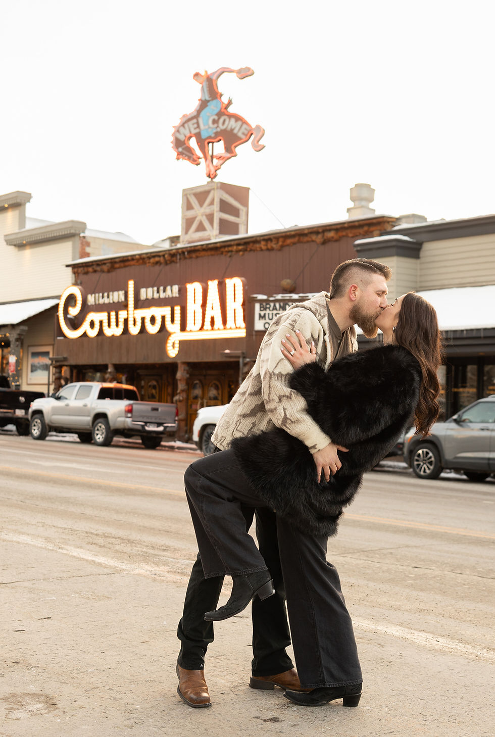 A couple kisses passionately outside the Million Dollar Cowboy Bar, with bright lights and cars in the snowy street background.