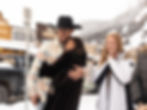 Three people outdoors in winter. Man in cowboy hat hugs a woman in a black coat. Woman in a white coat smiles. Snowy town setting.